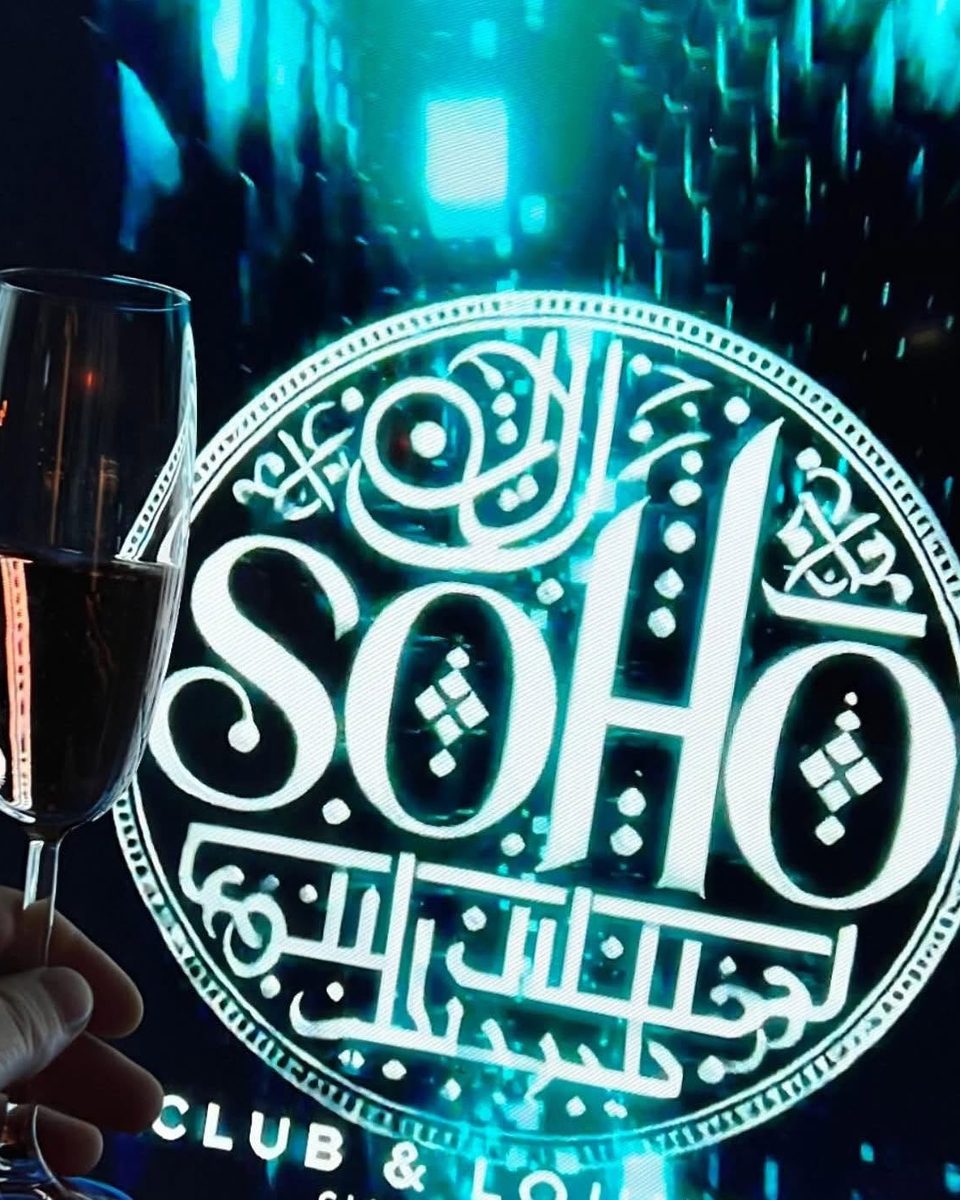 Pahar de șampanie ridicat în fața logo-ului SOHO de pe ecranul clubului